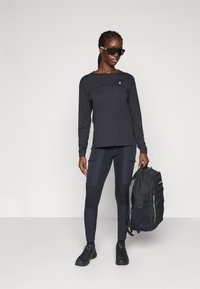 Black long-sleeve top with a curved seam, fitted black leggings with textured panels, and a dark backpack, worn with black shoes and sunglasses.