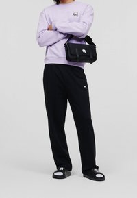 Persoon in een lavendelkleurig shirt met lange mouwen, zwarte broek, zwarte sandaal met witte sokken en een zwarte schoudertas, allemaal met een wit logo.