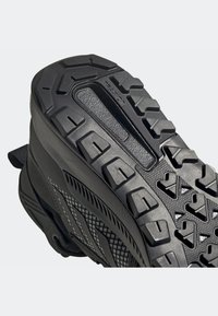 Černá sportovní obuv s texturovanou gumovou podrážkou, která má hluboké šlápnutí a geometrické vzory pro lepší přilnavost. Na bocích jsou síťované detaily a elegantní design.