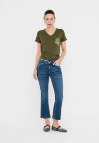 Olivegrünes V-Ausschnitt-T-Shirt mit einem Löwenmotiv auf der linken Brust, kombiniert mit blauen Schlagjeans und grauen Ballerinas. Ein einfaches, lässiges Outfit.