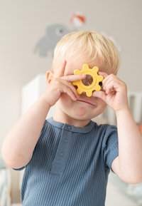 Geel, tandwielvormig speelgoed met een doorzichtige kern, omhoog gehouden bij het gezicht. Het kind draagt een geribd blauw T-shirt met korte mouwen en meerdere knopen.