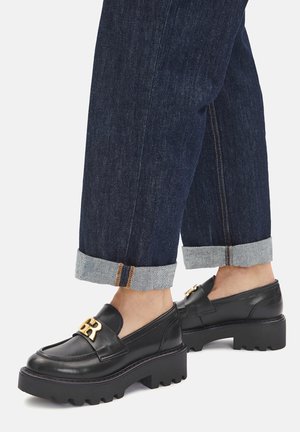 Czarne skórzane mokasyny z złotym akcentem logo, wyposażone w masywną podeszwę z bieżnikiem, zestawione z podwiniętymi ciemnymi dżinsami.