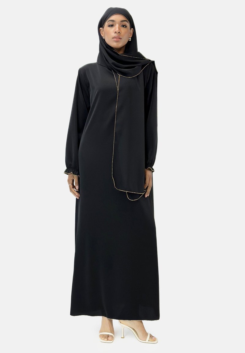 Robe noire à manches longues avec un col haut, ornée d'un détail doré à l'avant. Assortie à une écharpe assortie et des sandales à talons beiges.