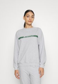 Šviesiai pilka džemperis su apvalia iškirpte, su žaliu "LACOSTE" logotipu ir akcentinėmis juostelėmis. Pagaminta iš švelnios, tekstūruotos medžiagos. Ilgos rankovės.
