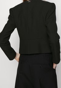 Černý zkrácený blazer se strukturovanými rameny, tvarovanými švy a hladkou texturou. Obsahuje malé logo na zadní straně a formální designové prvky.