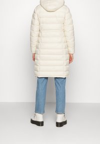 Długi, puszysty płaszcz w kolorze kremowym z kapturem, pikowana konstrukcja i elastyczne mankiety. Nosi się go na jasnoniebieskie dżinsy i białe buty z grubą podeszwą.