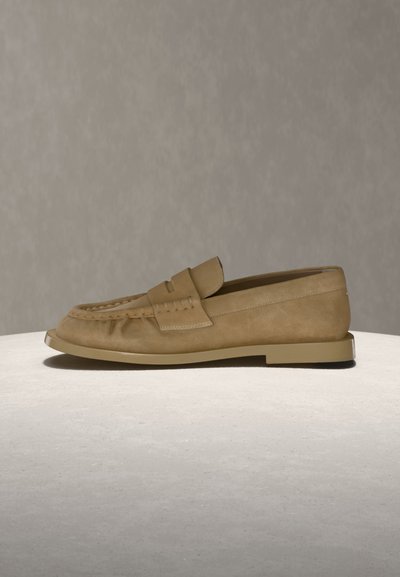 Mocassin pour homme en daim beige avec des détails cousus et une semelle plate beige, présenté sur un fond neutre sur une surface claire.