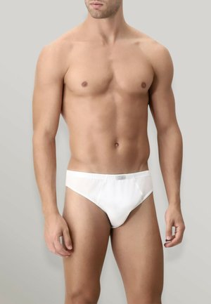 Uomo muscoloso indossa slip semplici in cotone bianco, in piedi contro uno sfondo grigio chiaro con le braccia rilassate lungo i fianchi.