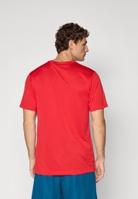 Rød kortærmet sportstrøje med en glat tekstur, rund hals og en afslappet pasform, vist fra bagsiden. Blå shorts er delvist synlige.