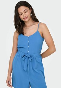 Mono azul sin mangas con escote en V, botones en la parte delantera y cintura atada. Hecho de una tela suave, presenta un corte holgado y una textura suave.