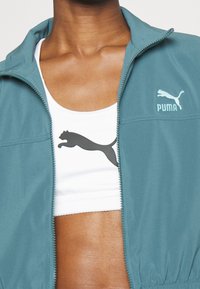 Teal løbejakke med lynlås og opretstående krave; hvid sports-bh med sort Puma-logo. Glat tekstur, moderne pasform, uden synlige beslag.