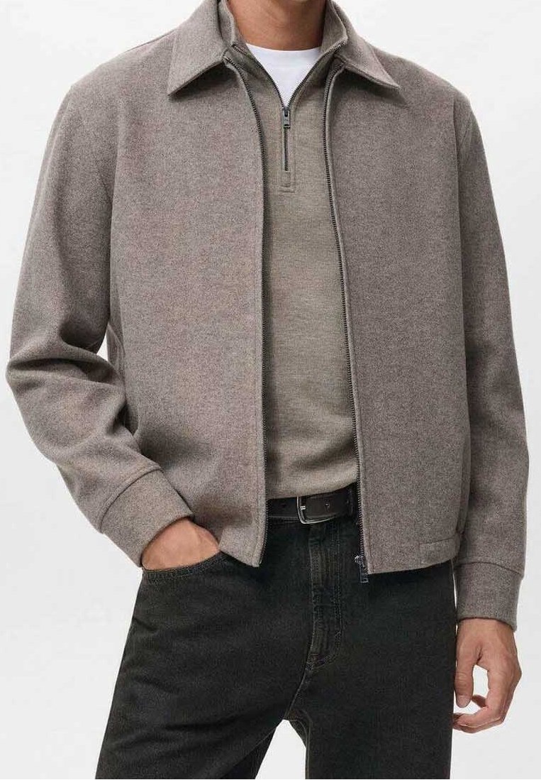 Homme portant une veste zippée marron clair par-dessus un pull beige avec fermeture éclair, ceinture noire et jean noir, une main dans la poche.