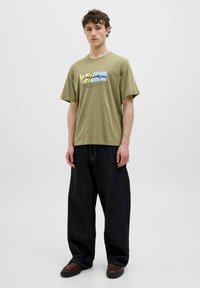 Olivenfarvet T-shirt med korte ærmer, rund halsudskæring og grafisk tryk med tekst og en blå accent. Båret med løse sorte jeans.