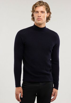 Joven con cabello rubio rizado, vestido con un suéter negro ajustado de manga larga y textura, y pantalones negros, de pie frente a un fondo liso.