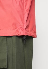 Červená nepromokavá bunda s elastickým lemem a nastavitelným stahovacím páskem. Kombinována s olivově zelenými kalhotami s plochými kapsami.