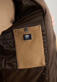 Giacca isolante marrone con interno che presenta una tasca in lana beige con etichetta cucita. La fodera è liscia e lucida, in contrasto con la texture.
