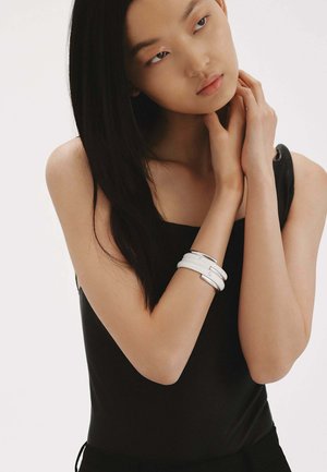 Top negro sin mangas, de tela suave. Pulseras blancas en capas con acentos plateados en una muñeca. Cabello negro liso, maquillaje natural.