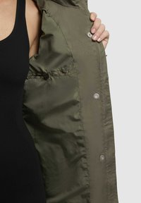 Veste vert olive avec une texture lisse et brillante, dotée de boutons-pression et d'une poche intérieure, partiellement ouverte pour révéler la doublure intérieure.