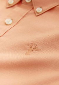 Polo color pesca con una finitura strutturata, caratterizzata da un piccolo ricamo a forma di aragosta e bottoni bianchi sul colletto.