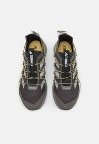 Scarpe da ginnastica grigie con accenti gialli, pannelli in rete e superfici testurizzate. Presentano lacci elastici e un marchio ben visibile sulla linguetta.