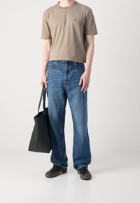 Bežové tričko s jemným logem, modré nohavice širokého střihu, černá taška přes rameno a tmavé tenisky se světlými podrážkami. Jednoduchý, neformální outfit.