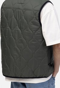 Gilet matelassé vert avec une surface texturée, un design rembourré et des bordures noires, porté par-dessus une chemise blanche et un jean bleu clair.