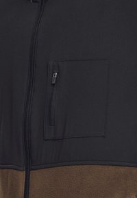 Svart og brunt glidelåsjakke med en teksturert fleece nederst, en glatt overdel og en glidelås lommesekk på brystet.