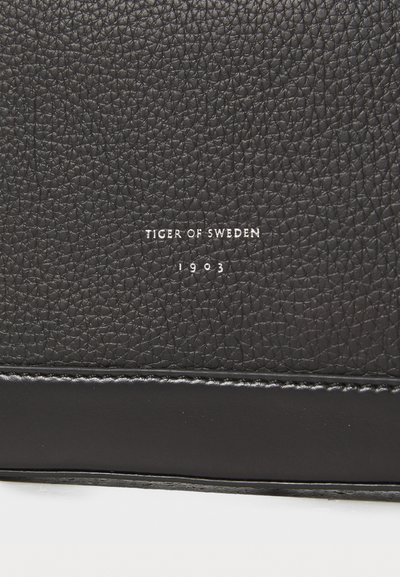 Musta tekstuuripinta nahkaa, jossa valkoinen "Tiger of Sweden 1903" logo on kohokuvioitu sujuvan nahkaompeleen ylle.
