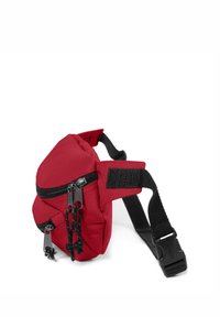 Červená látková ledvinka s dvojitými zipy, nastavitelným černým popruhem a přední nášivkou. Má kompaktní, šikmý design a texturovaný povrch.
