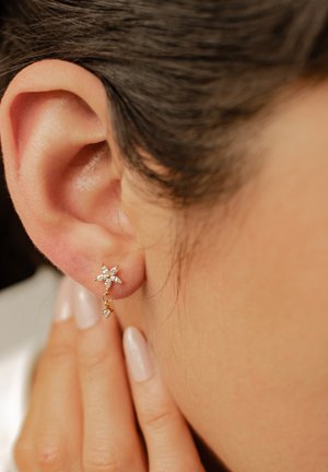 Nahaufnahme eines Ohrs mit kurzem braunem Haar, das einen kleinen goldenen, blumenförmigen Ohrring mit weißen Steinen trägt, Hand berührt den Hals mit blass lackierten Nägeln.