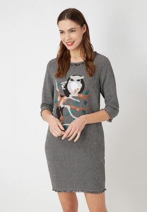 Mujer joven sonriendo, vistiendo un vestido de espiga en blanco y negro con mangas de 3/4 y un búho y una rama bordados en la parte delantera.