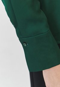 Maglietta a maniche lunghe di tessuto verde e liscio con polsini caratterizzati da un dettaglio di bottone nero e cuciture sottili lungo la cucitura.