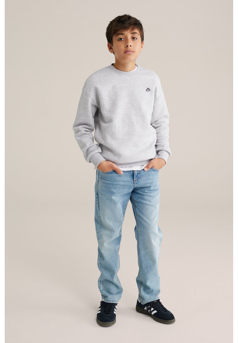 Junge steht mit den Händen in den Jeans-Taschen, trägt einen hellgrauen Pullover und blaue Jeans, blickt geradeaus vor neutralem Hintergrund.