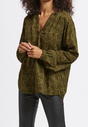 Mujer con blusa holgada de color verde oliva con estampado, escote en V pronunciado y mangas largas abullonadas, combinada con pantalones negros ajustados.