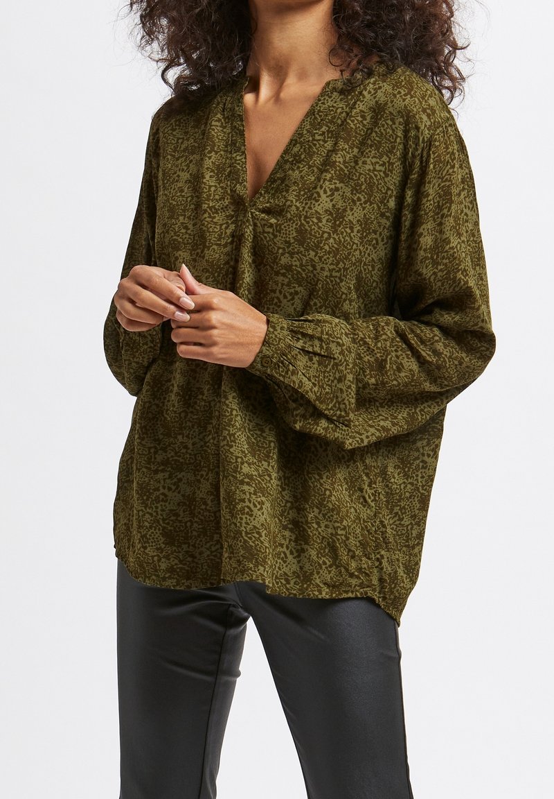 Femme portant un chemisier ample vert olive à motifs avec un profond décolleté en V et de longues manches bouffantes, associé à un pantalon noir ajusté.