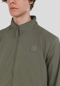 Lätt, olivgrön jacka med hög krage, dragkedjestängning och ett fyrkantigt logopatch på bröstet. Slät tygstruktur.