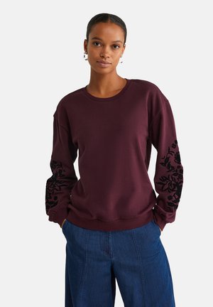 Jeune femme avec les cheveux tirés en arrière, portant un sweat à capuche pourpre foncé avec des motifs floraux noirs sur les manches et un pantalon en denim bleu lâche.
