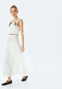 Outfit deux pièces texturé blanc comprenant un top court avec des accents de nœuds noirs et une jupe longue en couches. Des ballerines noires sont portées.