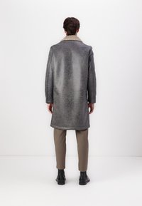 Largo abrigo gris hecho de material texturizado, con un gran cuello beige. Combinado con pantalones beige y botas negras. Visto desde atrás.