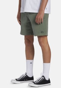 Olijfgroene korte broek voor heren, gemaakt van stevig materiaal, met een rechte snit, zijzakken en een klein logo-embleem. Draagt met zwarte sneakers en witte sokken.