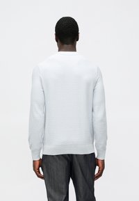 Gaiši pelēks džemperis ar ribotu tekstūru, garām piedurknēm un apaļu kakla izgriezumu. Kombinēts ar tumši pelēkām, viegli rakstainām biksēm.