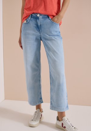 Person iført lyseblå afkortede jeans, hvide sneakers med rødt detaljer og en rød skjorte stoppet ind med den ene hånd i lommen.