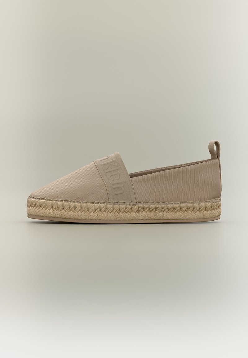 Espadrille beige à enfiler avec une semelle en jute tissé, bande élastique sur le dessus avec le nom de la marque embossé, et une languette en tissu au talon.
