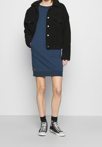 Černá fleecová krátká bunda s knoflíkovým zapínáním, která je kombinována s modrými šaty ke kolenům. Modelka má na sobě černé vysoké tenisky.