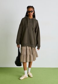Felpa oversize verde oliva con cappuccio e cordini, indossata sopra una gonna a strati leggera a quadri. Gli accessori includono occhiali da sole neri e una piccola borsa nera.