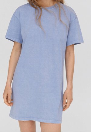 Femme portant une robe ample à manches courtes bleu clair, debout les bras détendus le long du corps, devant un fond uni.