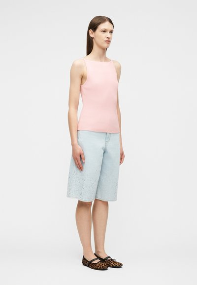 Γυναίκα που στέκεται φορώντας ένα ροζ αμάνικο τοπ, ανοιχτό μπλε σορτς μέχρι το γόνατο με πιτσιλιές μπογιάς, και μπαλαρίνες με λεοπάρ σχέδιο και μαύρες φιόγκους.