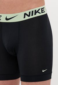 Črne bokserice z lahnim zelenim pasom z logotipom Nike, ki imajo gladko teksturo in oprijet kroj za udobje in podporo.