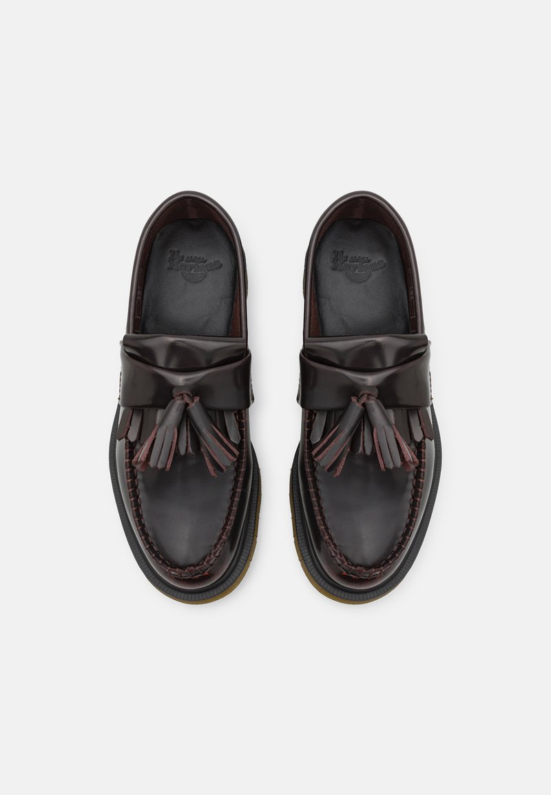 Braune Lederlatschen mit einer Quastenakzent, Mokassinnaht und einer Gummisohle. Das Innere ist dunkel und zeigt ein Markenlogo.