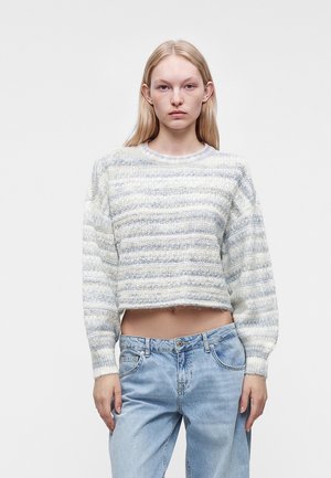 Jeune femme aux cheveux blonds et raides portant un pull court léger rayé et un jean bleu clair ample, debout devant un fond uni.